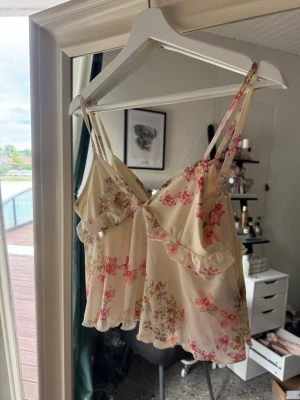 Blommig festtopp/pyjamas med volang - Söt loungewear-top i ljusbeige med rosa och röda blommor. Tunt, lätt material med volangdetaljer och smala axelband. Perfekt för en avslappnad dag hemma eller fest. Luftig och feminin design med romantisk känsla.
