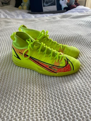 Nike Mercurial gula inne fotbollsskor - Säljer ett par Nike Mercurial inne fotbollsskor i neon-gult med svarta och röda detaljer. Skorna har hög krage för extra stöd runt ankeln och är tillverkade i ett lätt syntetmaterial. Snygg design med stora Nike-loggor på sidorna och snörning framtill.