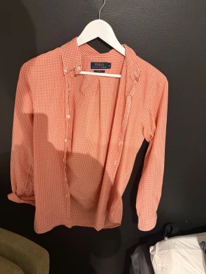 Rutig orange skjorta från Polo Ralph Lauren - Säljer en snygg orange och vit rutig skjorta från Polo Ralph Lauren. Skjortan har klassisk krage, långa ärmar och knappar framtill. Perfekt för dig som gillar en avslappnad men ändå stilren look.