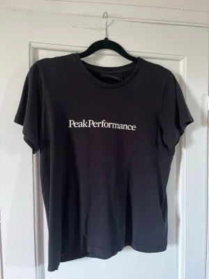 Svart Peak Performance t-shirt - Klassisk svart t-shirt från Peak Performance med vit logga tryckt på bröstet. T-shirten har rund halsringning och korta ärmar. Tillverkad i mjuk bomull för en skön och avslappnad stil.