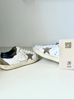 Golden Goose vita sneakers med stjärna - Golden Goose sneakers i vitt läder med grå mockadetaljer och ikonisk stjärna på sidan. Skorna har mörkblå hälkappa, vita snören och vintageinspirerad sula. Perfekta för en avslappnad och trendig look. Har fått lite rosa färg på sig  men man tänker knappt på det vid användning. Äkthetsbeviset följer ej med