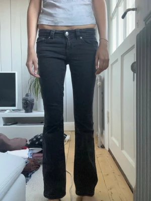 Svarta bootcut jeans med fickdetalj - Snygga svarta bootcut jeans med låg midja och en unik fickdetalj med knapp framtill. Jeansen har klassisk ficksdesign och är tillverkade i ett stretchigt denimtyg som sitter bekvämt. Perfekta för dig som gillar en tidlös och clean look.