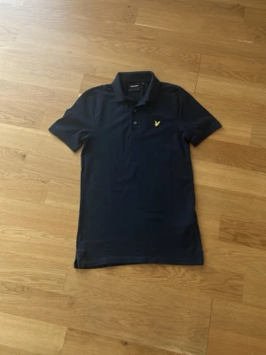 Mörkblå piké med krage - Snygg mörkblå pikétröja med klassisk krage och korta ärmar. Tröjan har knappar framtill och en gul broderad logga på bröstet. Perfekt till jeans eller shorts för en clean och stilren look.