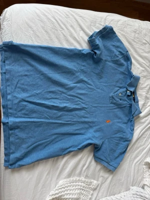Blå pikétröja från Polo Ralph Lauren - Säljer en blå pikétröja från Polo Ralph Lauren i slim fit. Tröjan har klassisk krage, korta ärmar och den ikoniska broderade loggan i orange på bröstet. Perfekt till jeans eller shorts för en clean och stilren look. Storlek M
