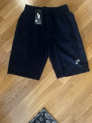 Svarta shorts från Nike med snörning - Svarta shorts från Nike med elastisk midja och snörning. Klassisk logga i vitt på ena benet och två sidofickor. Perfekta för en sportig och avslappnad stil.