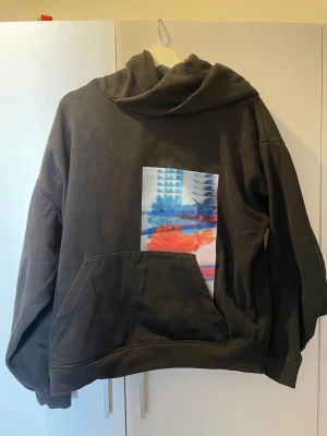 Svart hoodie från Calvin Klein Jeans - Säljer en svart hoodie från Calvin Klein Jeans med stor huva och magficka. På framsidan finns en färgglad abstrakt patch i blått, rött och vitt. Baksidan har ett vitt tryck med texten 'Calvin Klein Jeans NEW YORK'. Perfekt för en chill och trendig look.