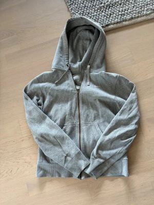 Grå Nike zip hoodie med huva - Säljer en Nike stilren grå hoodie med dragkedja framtill och stor huva. Hoodien har ribbade muddar vid ärmslut och nederkant samt två fickor framtill. Perfekt för en avslappnad och bekväm stil. Tröjan har en liten defekt på sista bilden men den är inte märkbar. 