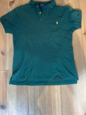 Grön pikétröja från Polo Ralph Lauren - Klassisk grön pikétröja från Polo Ralph Lauren med gul broderad logga på bröstet. Tröjan har korta ärmar, krage och knappar vid halsen. Slim fit-modell i mjuk bomull som passar perfekt till jeans eller shorts.
