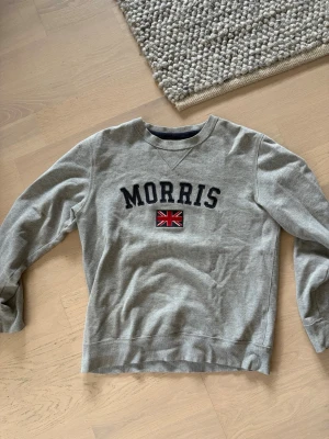 Grå sweatshirt från Morris - Snygg grå sweatshirt från Morris med broderad text och brittisk flagga på bröstet. Tröjan har rund halsringning, långa ärmar och ribbade muddar. Perfekt för en avslappnad och stilren look. Den är i storlek M.