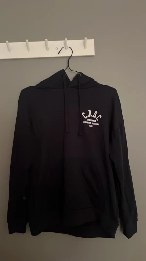 Marinblå hoodie med C.A.S.C tryck - Marinblå hoodie med huva och dragsnören. På bröstet och ryggen finns vitt tryck med texten 'C.A.S.C California Athletics & Social Club'. Klassisk passform och mjukt material, perfekt för en avslappnad stil.