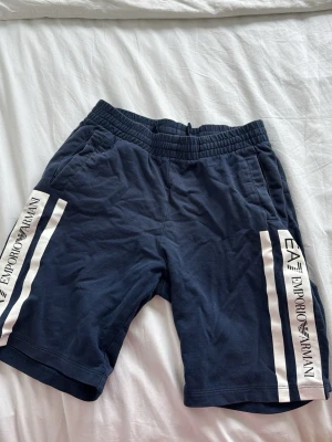 Marinblå shorts från Emporio Armani - Snygga marinblå shorts från Emporio Armani med vita sidopaneler och logga längs båda benen. Shortsen har elastisk midja och är perfekta för en avslappnad och sportig stil. Tillverkade i mjukt material för extra komfort.