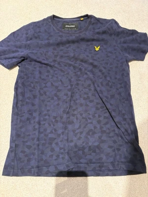 Blå mönstrad t-shirt från Lyle & Scott - Snygg blå t-shirt från Lyle & Scott med diskret geometriskt mönster och gul logga på bröstet. Klassisk rund hals och korta ärmar. Perfekt till jeans eller shorts för en avslappnad stil.