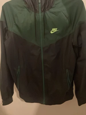 Grön och svart Nike vindjacka - Snygg vindjacka från Nike i mörkgrönt och svart med dragkedja framtill och huva. Jackan har två fickor med dragkedja och en klassisk Nike-logga i ljusgrönt på bröstet. Perfekt för blåsiga dagar och sportiga outfits.