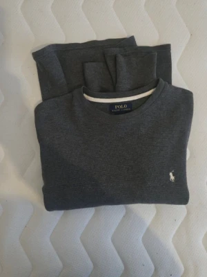 Grå tröja från Polo Ralph Lauren - Snygg grå tröja från Polo Ralph Lauren i 100% bomull. Klassisk rund halsringning med vit detalj och den ikoniska broderade loggan på bröstet. Perfekt för en stilren och avslappnad look.