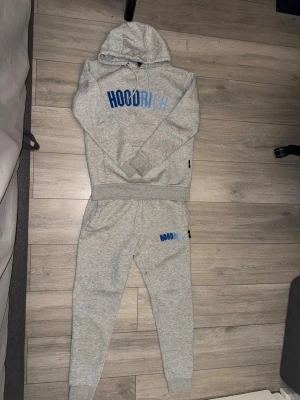 Gråa mjukisbyxor från Hoodrich+ Hoodien - Säljer ett par gråa mjukisbyxor+ hoodie från Hoodrich med blå logga på vänster lår. Byxorna har resår i midjan och vid bensluten för en skön passform. Perfekta för chill dagar och streetwear-stil. Pris kan diskuteras vid snabb affär