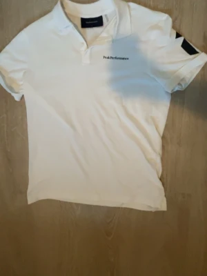 Vit pikétröja från Peak Performance - Klassisk vit pikétröja från Peak Performance med liten logga på bröstet. Tröjan har krage, knappar vid halsen och korta ärmar. Perfekt till både jeans och shorts för en clean och sportig look.