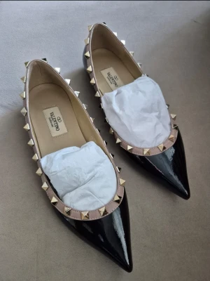 Valentino svarta lackade ballerinaskor - Säljer ett par svarta ballerinaskor från Valentino i lackat skinn med spetsig tå. Skorna har beige kant med guldnitar runt öppningen som ger en edgy look. Perfekta för dig som vill ha något extra till outfiten.