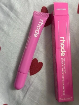 Rhode Peptide Lip Tint Shortcake - Säljer ett Rhode Peptide Lip Tint i färgen Shortcake. Läppbalsamet kommer i en rosa tub med matchande förpackning och har en smidig applikator. Innehåller 10 ml och ger en lätt rosa ton till läpparna. Perfekt för dig som vill ha återfuktade och snygga läppar.