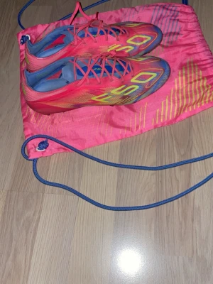 Färgglada fotbollsskor F50 - Säljer ett par riktigt snygga fotbollsskor i modellen F50. Skorna är rosa med gula och blå detaljer samt blå insida. De har snörning och en aerodynamisk form som passar perfekt för snabba spelare. Kommer med en matchande rosa gympåse med blå snören.