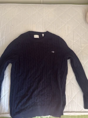 Mörkblå kabelstickad tröja GANT - Mörkblå kabelstickad tröja från GANT med rund hals och ribbade muddar. Tröjan har ett diskret broderat GANT-emblem på bröstet och är tillverkad i ett mjukt stickat material. Perfekt för dig som gillar klassisk och stilren design.