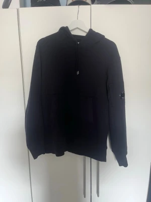Marinblå hoodie från C.P. Company - Säljer en äkta marinblå hoodie från C.P. Company med klassisk huva och dragsko. Tröjan har en ficka på magen och den ikoniska linsen på ärmen. Tillverkad i mjukt bomullsmaterial som är skönt att bära. Perfekt för dig som gillar streetwear och stilrena plagg.