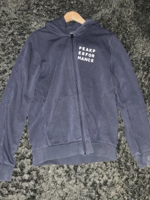 Mörkblå hoodie Peak Performance - Snygg mörkblå hoodie från Peak Performance med dragkedja framtill och vit logga på bröstet. Klassisk huva och två fickor framtill. Perfekt för en avslappnad och sportig stil. Tillverkad i mjukt material för extra komfort.