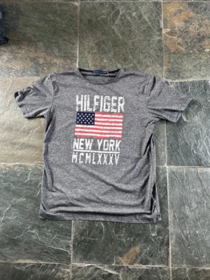 Grå Hilfiger t-shirt med USA-flagga - Snygg grå t-shirt från Hilfiger med tryck av amerikanska flaggan och texten 'NEW YORK MCMLXXXV' på bröstet. T-shirten har rund hals och korta ärmar, perfekt för en avslappnad streetstyle-look.