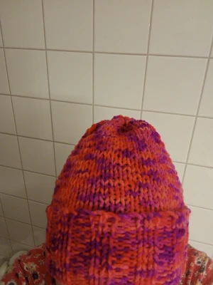 Stickad mössa i rosa och lila - Säljer en handstickad mössa i en mix av rosa och lila nyanser. Mössan har en uppvikt kant och är gjord i ett tjockt, mjukt garn som håller dig varm. Perfekt för kalla dagar och ger en färgklick till din outfit. Material Akrylic och handtvätt 30 grader.(Går att fås med tofs om man skulle önska detta).