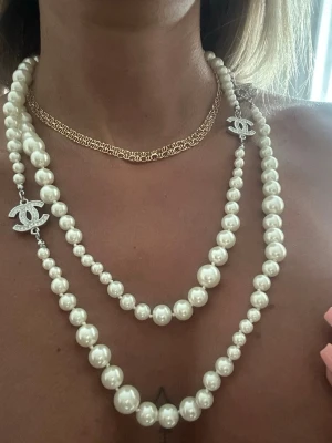 Chanel pärlhalsband med logga - Lyxigt Chanel halsband med vita pärlor i två rader och silverfärgade detaljer. Halsbandet har två ikoniska Chanel-loggor med gnistrande stenar. Perfekt statement piece för dig som vill sticka ut och addera exklusiv känsla till din outfit.