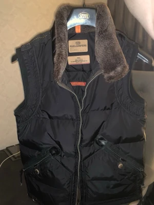 Svart dunväst från Parajumpers - Snygg svart dunväst från Parajumpers med mjuk, brun fuskpälskrage och två stora fickor med dragkedja och tryckknapp. Västen har en sportig look med quiltad design och dragkedja framtill. Perfekt för lager på lager under kyliga dagar. Notera att backsidan har rivmärken men inget betydligt synligt. Inköpspris va 4000 säljer för 3000 seriösa köpare kan diskuteras med
