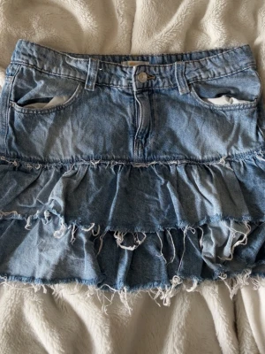 Blå jeanskjol med volanger - Säljer en blå jeanskjol med två lager volanger och råa, fransiga kanter. Kjolen har klassiska fickor fram och bak samt bälteshällor. Materialet är denim i en ljusblå tvätt som ger en avslappnad och trendig look.