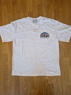 Vit t-shirt från Casablanca - Säljer en vit t-shirt från Casablanca med ett färgglatt tryck av en byggnad och logga på bröstet. Klassisk rund hals och korta ärmar. Tillverkad i mjuk bomull som känns skön mot huden. Perfekt för dig som gillar stilrena plagg med en twist.