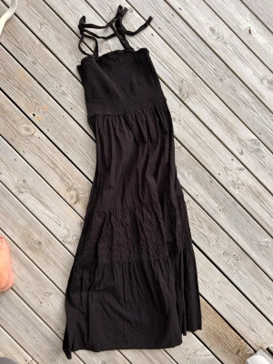 Svart långklänning Pernilla Wahlgren - Svart långklänning från Pernilla Wahlgren med smock upptill och knytbara axelband. Klänningen har en luftig passform och flera volanglager som ger ett snyggt fall. Perfekt för varma dagar och enkel att styla med accessoarer.