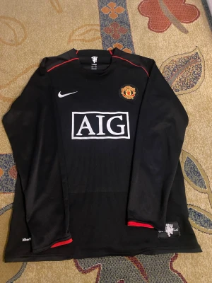 Manchester United Ronaldo tröja svart - Snygg svart långärmad Manchester United matchtröja från Nike med AIG-logga på bröstet och Ronaldo 7 på ryggen. Tröjan har röda detaljer vid axlar och ärmslut samt klubbmärke på bröstet. Perfekt för fotbollsfans och samlare. Passar både XL och L.