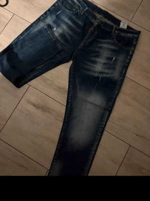 Blå slitna jeans från Dsquared2 - Säljer ett par blå jeans från Dsquared2 med slitningar och tvättade detaljer. Jeansen har klassisk femficksmodell, raka ben och tydliga slitningar framtill. Materialet är denim och färgen är mörkblå med ljusare partier. Perfekta för en avslappnad och trendig look.