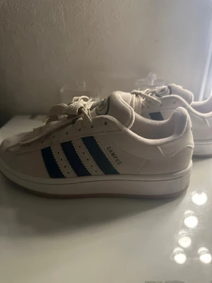 Adidas Campus beige/blå sneakers - Snygga äkta Adidas Campus sneakers i beige mocka med blå detaljer. Perfekta skor om ni vill ha något bekvämt till vardags. Har andvända dessa skor 2-3 gånger. Sedan får ni med mörkblåa snören till skorna. Tips till skolstarten om ni vill ha något som ger er nått litet extra till outfiten. 