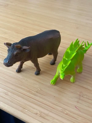 Plastdjur: ko och grön dinosaurie - Två plastfigurer, en brun ko med detaljerade drag och en ljusgrön dinosaurie med taggar längs ryggen. Perfekta som leksaker eller dekoration. Båda är gjorda i hårdplast och har realistiska former.