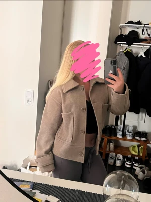 Beige ulljacka med stora fickor - Snygg beige jacka i ull med stora framfickor och bred krage. Jackan är croppad och har knappar framtill samt detaljer vid ärmsluten. Perfekt för höst och vår när du vill ha en stilren look.