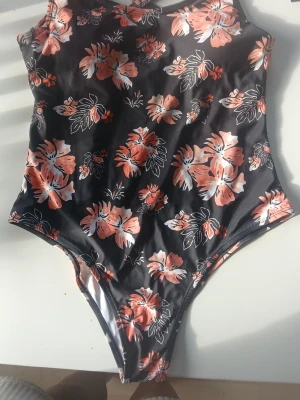 Svart baddräkt med blommigt mönster - Snygg svart baddräkt med rosa och vita blommor samt gröna blad. Modellen har smala axelband som korsas i ryggen för en trendig look. Materialet är stretchigt och följsamt, perfekt för strand eller pool. Blommigt mönster ger en fräsch och somrig känsla.