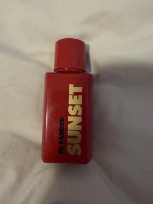 Jil Sander SUNSET parfym - Jil Sander SUNSET parfym i en snygg, rektangulär röd flaska med guldfärgad text. Flaskan har en matchande röd kork och modern design som sticker ut på hyllan. Perfekt för dig som gillar unika och färgstarka doftflaskor.