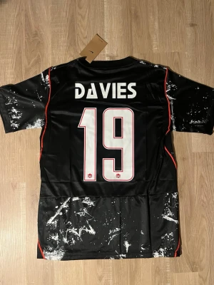 Svart Kanada fotbollströja Davies - Säljer en svart Kanada fotbollströja med vitt splattermönster, röd logga och röda detaljer. Nummer 19 och namnet Davies på ryggen. Tillverkad i lätt och ventilerande material, perfekt för träning eller match. HELT NY!