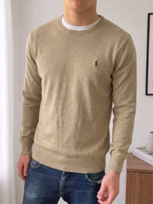 Beige tröja från Ralph Lauren - Klassisk beige tröja från Ralph Lauren med rund halsringning och diskret logga broderad på bröstet. Tröjan har ribbade muddar vid ärmslut och nederkant. Perfekt för en stilren och avslappnad look.