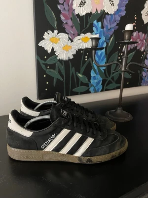 Adidas Spezial svarta sneakers - Säljer ett par Adidas Spezial sneakers i svart mocka med vita detaljer och klassiska tre ränder på sidan. Skorna har svart snörning, vit hälflik och gummisula med Adidas-logga. Perfekta för dig som gillar retrostil och streetwear.