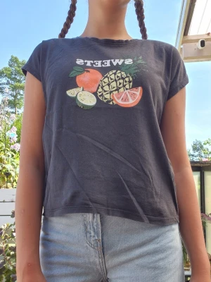Svart t-shirt med frukttryck - Cool svart t-shirt med färgglatt frukttryck och texten SWEETS på bröstet. T-shirten har korta ärmar och en avslappnad passform, perfekt till jeans eller shorts. Materialet känns mjukt och skönt mot huden.
