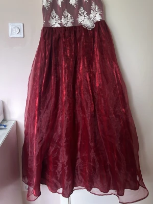 Vinröd långklänning med spetsdetaljer med fyra lager  - Elegant vinröd långklänning med glansig kjol och överdel i vitt med broderade spetsdetaljer och blommönster. Klänningen har ärmlös design och markerad midja, perfekt för speciella tillfällen.