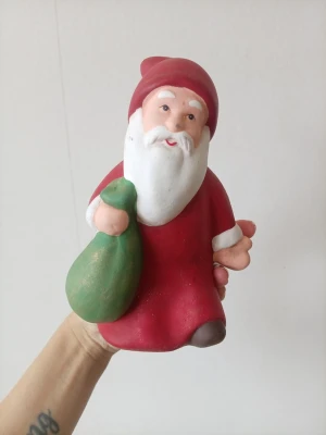 Tomtefigur i keramik med säck - En söt tomtefigur i keramik, målad i rött, vitt och grönt. Tomten har en klassisk röd luva, vit skägg och håller i en grön säck. Perfekt som dekoration till jul och att ställa på hyllan eller bordet. Färgen ha släppt på vissa ställen men funkar ändå 