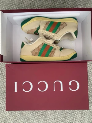 Gucci sneakers med gröna och orange detaljer - Snygga sneakers från Gucci i beige med gröna och orange ränder på sidan. Skorna har klassiskt GG-mönster i canvas, gröna detaljer vid sula och häl samt beige snörning. Perfekt för dig som vill sticka ut med exklusiva och trendiga sneakers.