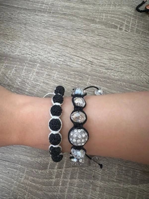 Två svarta armband med pärlor - Säljer två snygga armband med svarta och vita knytband. Ena armbandet har svarta pärlor med vita detaljer, det andra har glittriga pärlor och stora kristalliknande pärlor. Justerbara band för perfekt passform. Perfekt accessoar för att lyfta din stil.