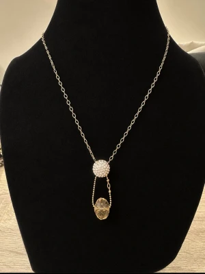 Guldhalsband med kristall detaljer - Snyggt halsband i guld med tunn kedja, dekorerat med en rund pärlliknande detalj och en droppformad klar kristall. Perfekt för dig som gillar stilrena och eleganta accessoarer med en unik twist.