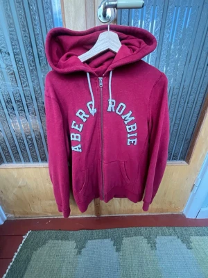 Röd hoodie från Abercrombie & Fitch - Snygg röd hoodie från Abercrombie & Fitch med dragkedja och stora vita bokstäver på bröstet. Hoodien har huva med dragsko och två fickor framtill. Perfekt för en avslappnad och cool stil.